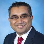 Smiling man in glasses wearing a suit and tie, with a light blue background.