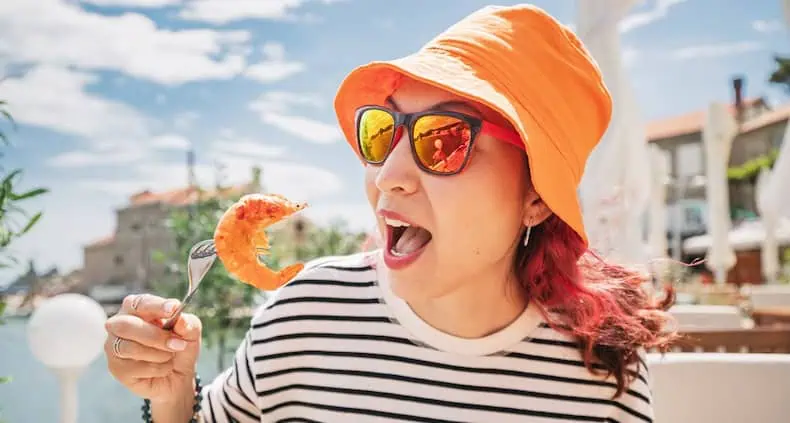 A woman in an orange hat and striped shirt eagerly holds a shrimp on a fork, smiling against a sunny outdoor backdrop.